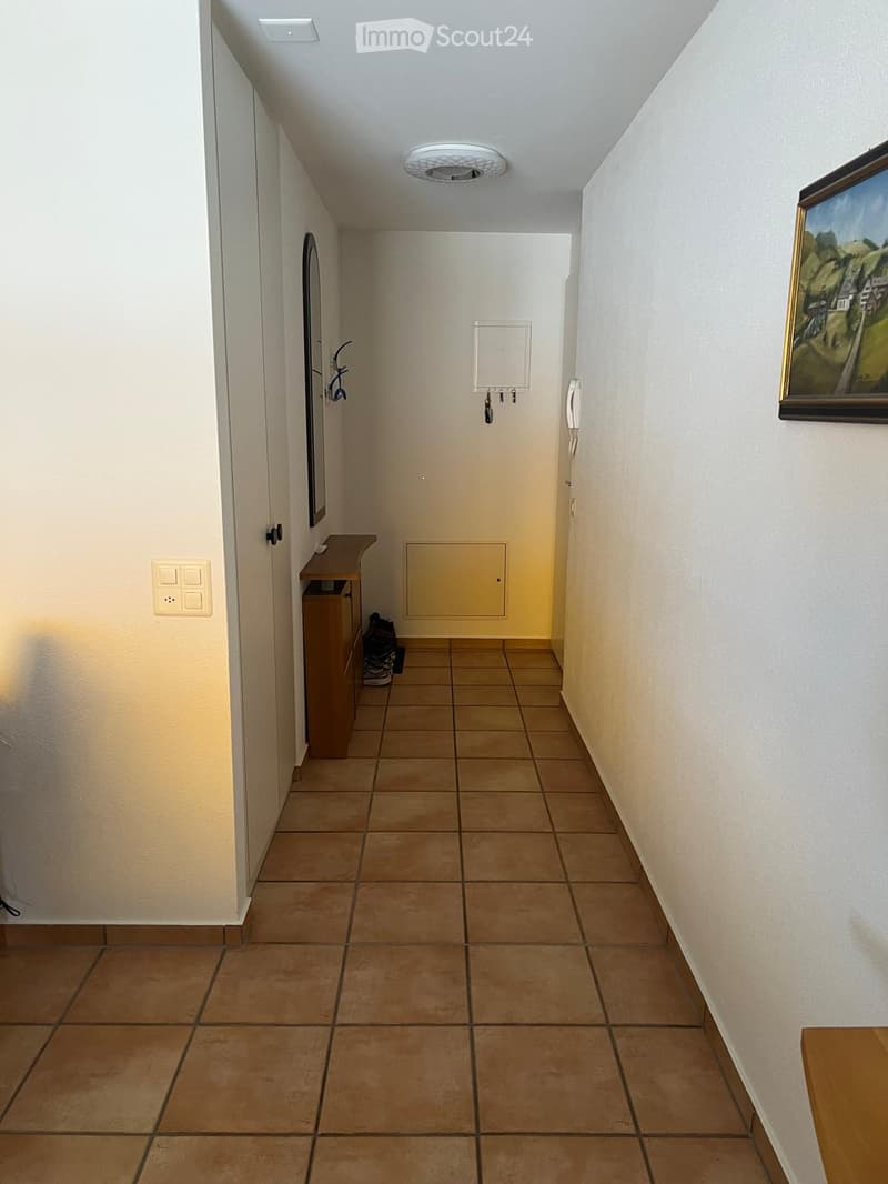 A narrow hallway with tiled flooring, white walls, and a small wooden bench on the left side. A painting hangs on the right wall, and a mirror is mounted on the left wall.