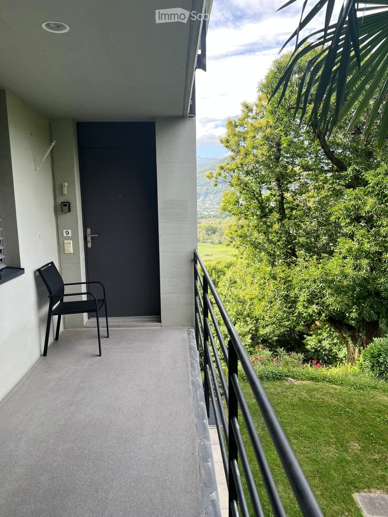 Balcony with a black chair, metal railing, and a door leading to the interior. The view includes a green garden and distant mountains.