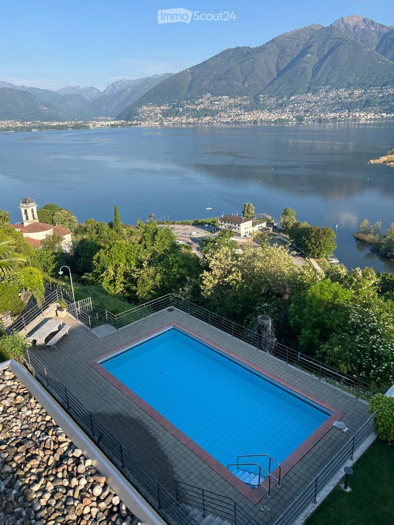 A rooftop terrace with a swimming pool overlooking a scenic lake and distant mountains, surrounded by greenery and a small seating area.