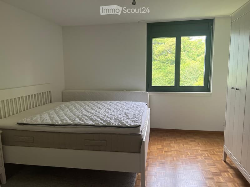 Bright bedroom with wooden flooring, large window, white bed with quilted mattress, and built-in wardrobe.