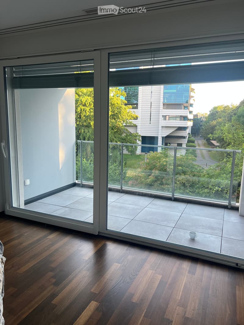 A modern, well-lit room with wooden flooring and large glass doors leading to a balcony. The balcony features a tiled floor and a glass railing, offering a view of greenery and modern buildings.