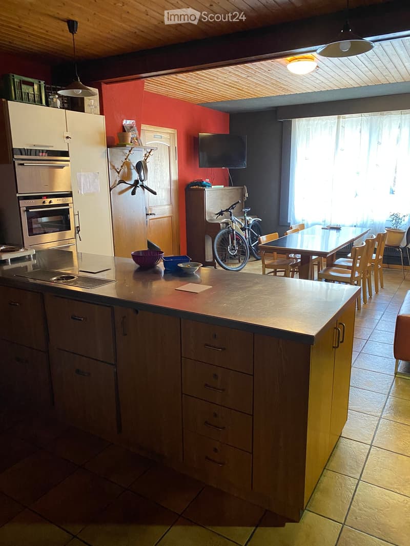 The kitchen features a central island with a wooden countertop, a refrigerator, an oven, and a screen mounted on the wall. A dining table with chairs is visible, along with a bicycle leaning against the wall and several bowls on the island.