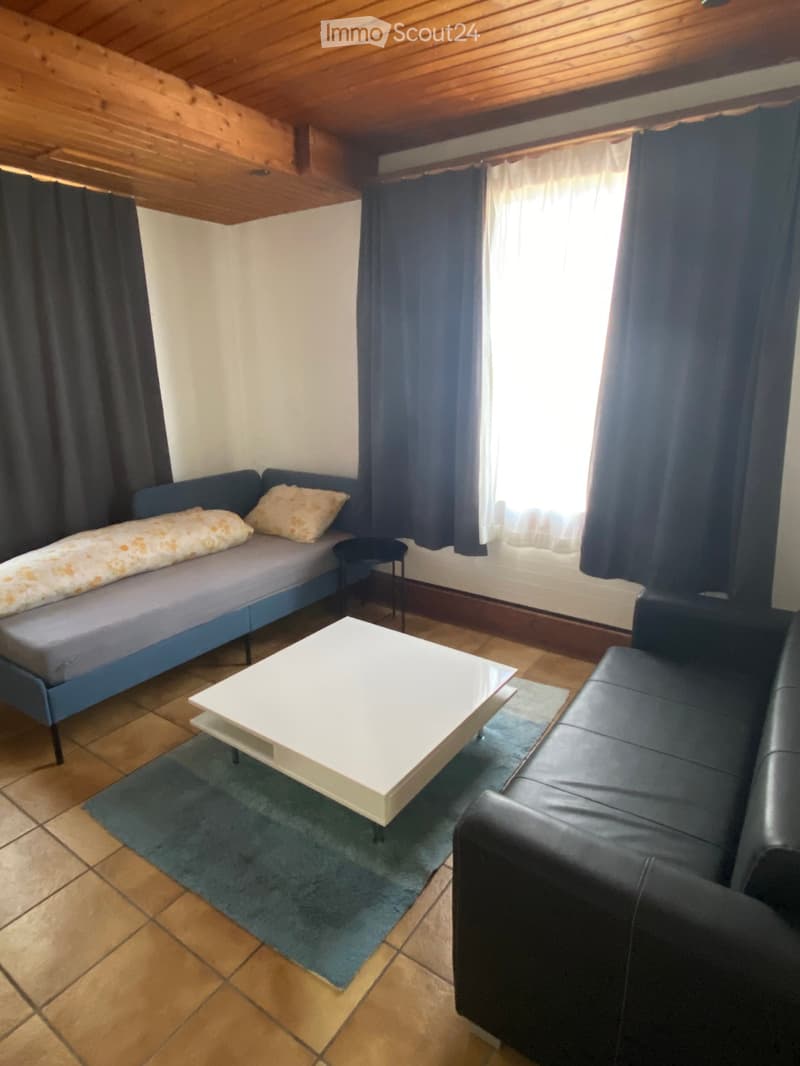 A small, cozy room with a blue sofa, a white coffee table, a bed with pillows, and a small stool near the window.