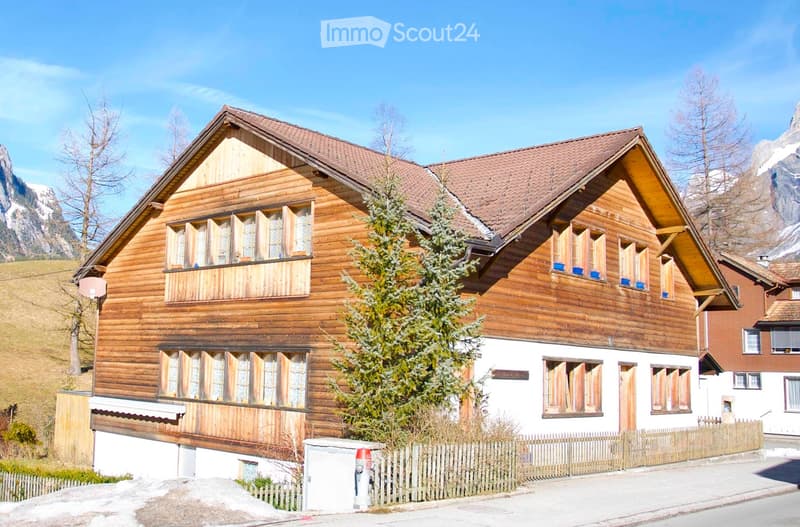 A two-story wooden house with a sloped roof, multiple windows, and a small balcony. The house is surrounded by a wooden fence and trees, with a scenic mountain view in the background.