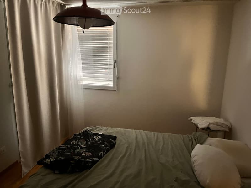 A small bedroom with a single bed, pillows, a hanging lamp, and a window with white curtains.