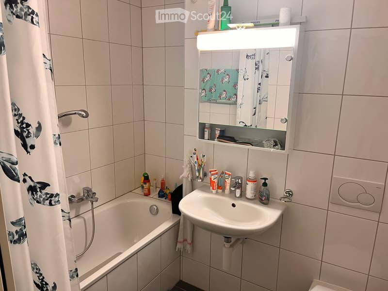 Bathroom with a bathtub, sink, mirror, and various toiletries. The walls are tiled, and there is a shower curtain with a decorative pattern.