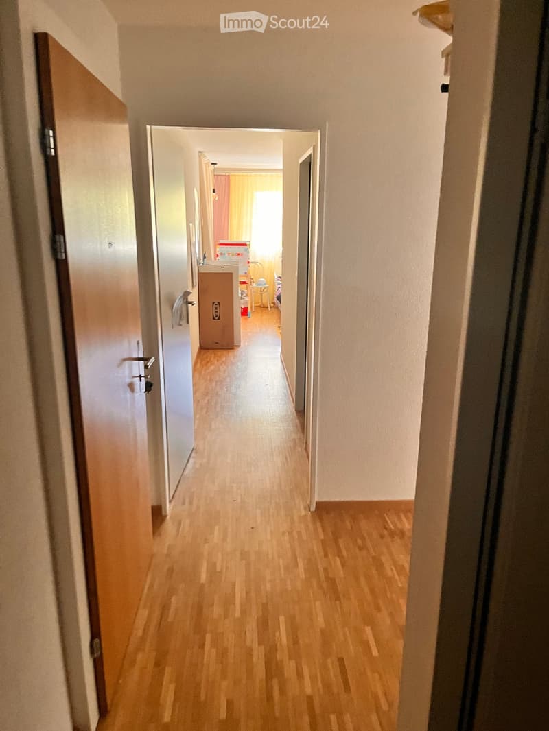 A narrow hallway with wooden flooring and white walls, leading to an open room with furniture and boxes visible at the end.