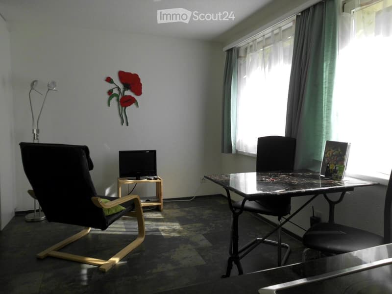 A furnished room with a desk, chair, TV, and a comfortable armchair near a window with curtains.