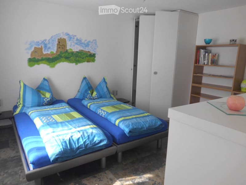 A well-lit bedroom with two single beds, blue and green bedding, a nightstand, a bookshelf, and a small table with a decorative item.