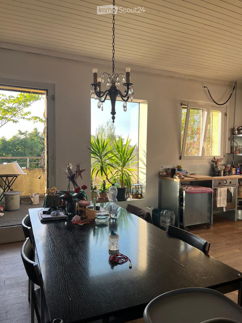 A modern, well-lit dining area with a large wooden table, chairs, and a chandelier. The space is connected to a kitchen area with appliances and a view of greenery outside.