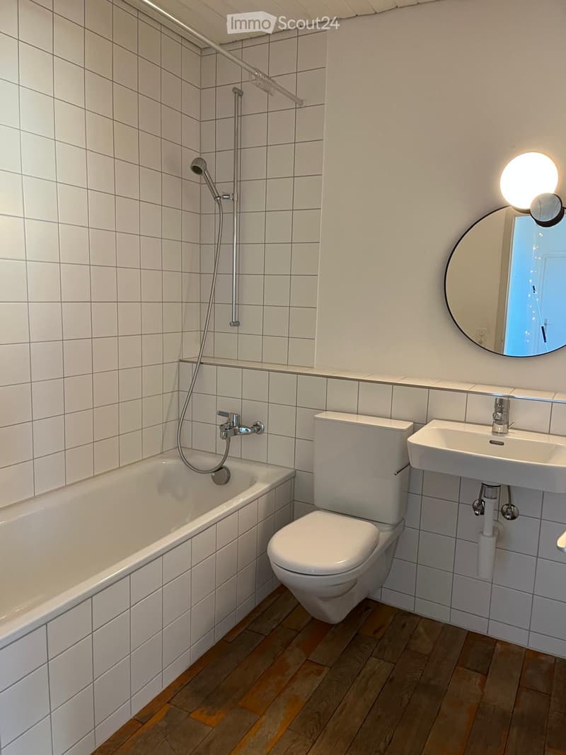 Modern bathroom with white tiled walls, a bathtub, toilet, sink, and a round mirror with a built-in light. The floor is wooden.