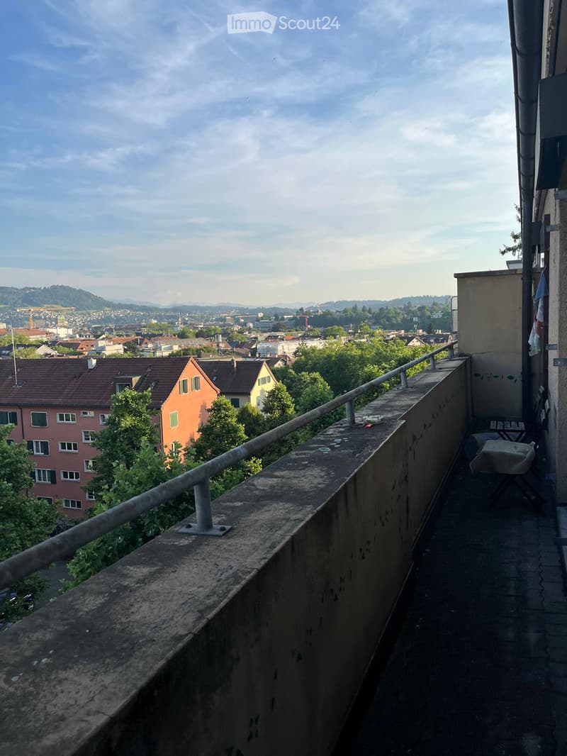 Balcony with metal railing, overlooking a cityscape with buildings, trees, and distant hills under a partly cloudy sky.