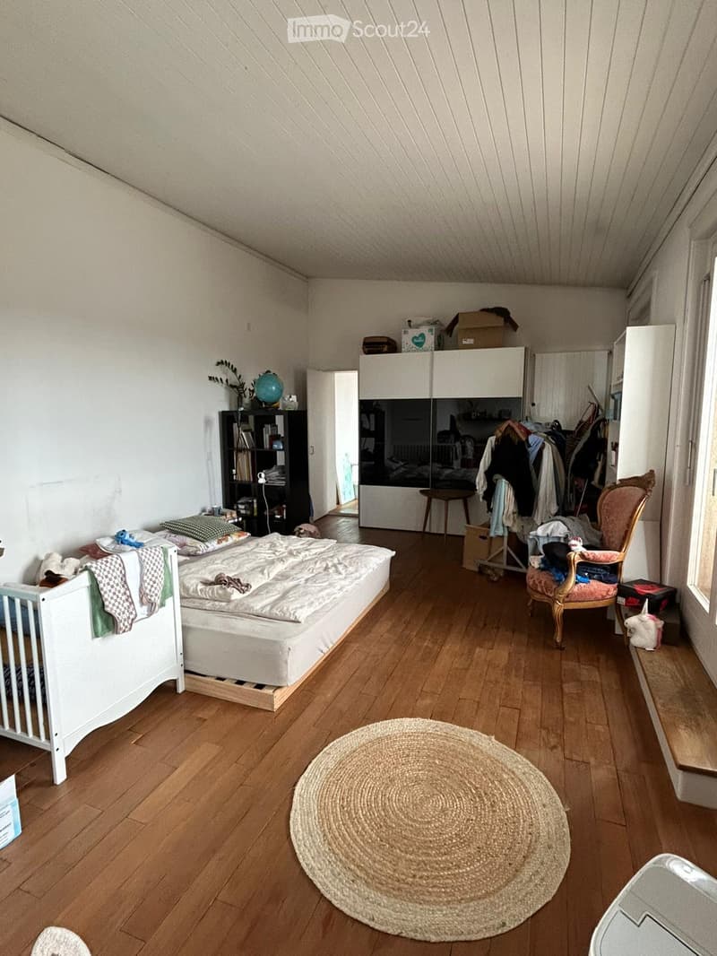 Cozy attic bedroom with wooden floors, slanted ceilings, a single bed, a round woven rug, a decorative chair, a bookshelf, and a wardrobe with an open closet area.