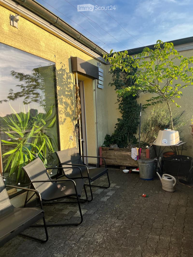 Outdoor patio with two black metal chairs, a small table, a potted plant, a watering can, and a trash can. The area is paved with stone tiles and has a view of a building with yellow walls and a glass door.