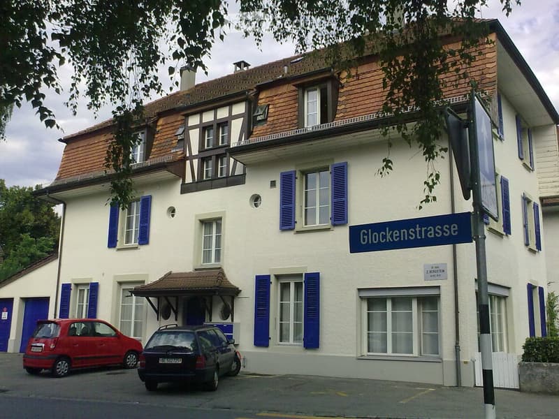 White building with brown roof, blue shutters, two cars parked in front