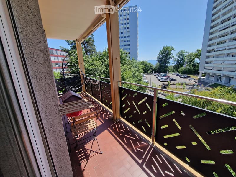 Balcony with wooden table and chairs, metal railing, and view of parking lot and buildings.
