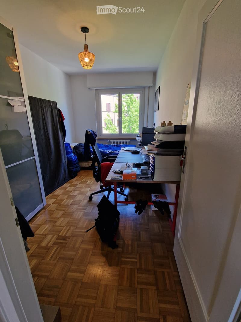 A furnished room with a wooden floor, a desk with a laptop, keyboard, and mouse, a chair, a bed with a curtain, and a hanging lamp.