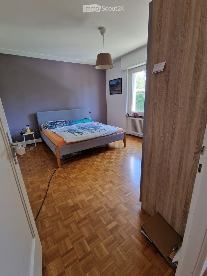 Bedroom with wooden herringbone flooring, grey headboard bed, nightstand, lamp, and picture frame on wall.