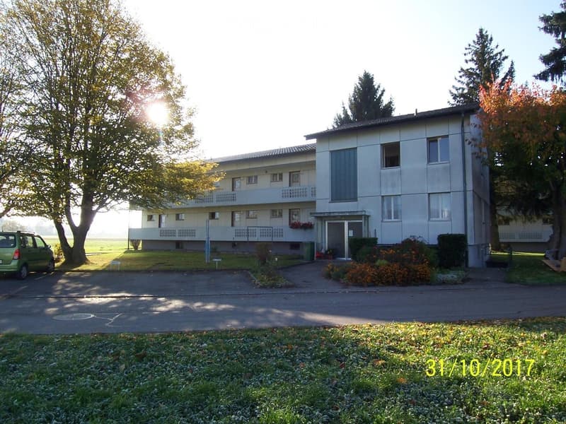 Two-story residential building with balconies, surrounded by trees and a paved driveway. Green car parked nearby.