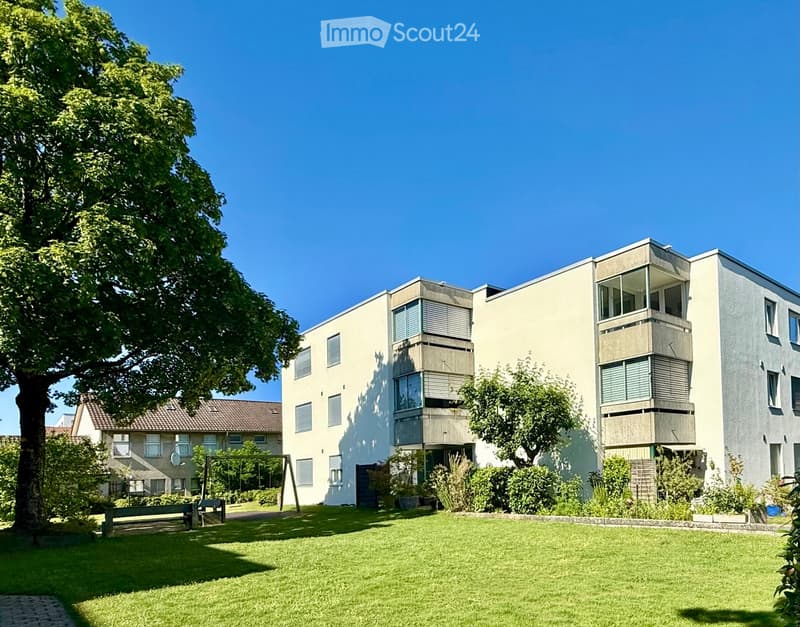 Modern multi-story residential building with balconies, surrounded by greenery and a lawn.