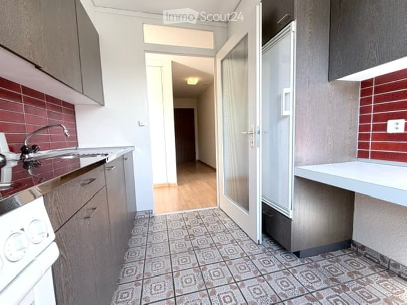 Modern kitchen with red brick backsplash, white cabinets, stainless steel sink, and stove. The floor features a patterned tile design, and the space is well-lit with natural light from the adjacent hallway.