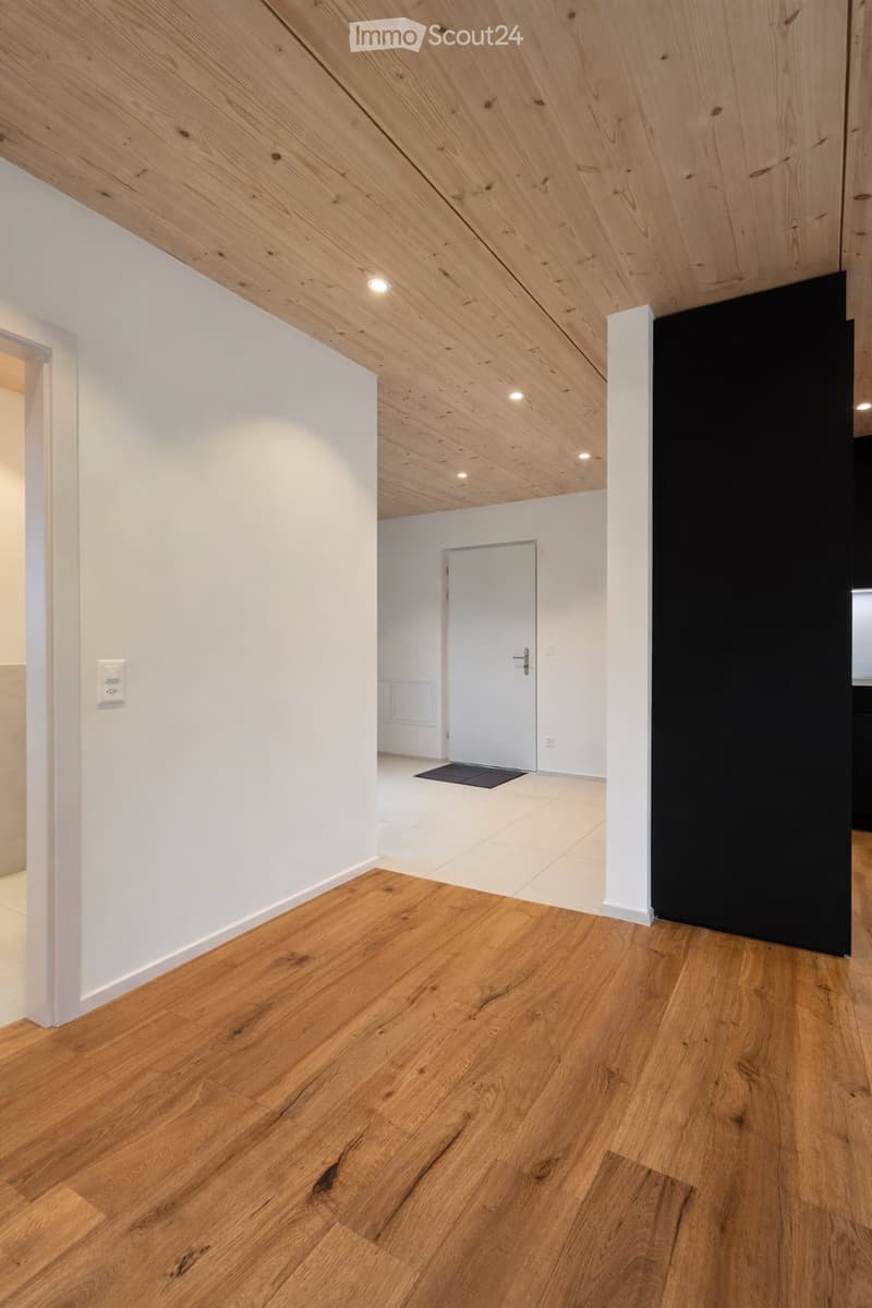 Empty room with wooden flooring, white walls, and a slanted wooden ceiling. A doorway leads to another room with white tiled flooring.