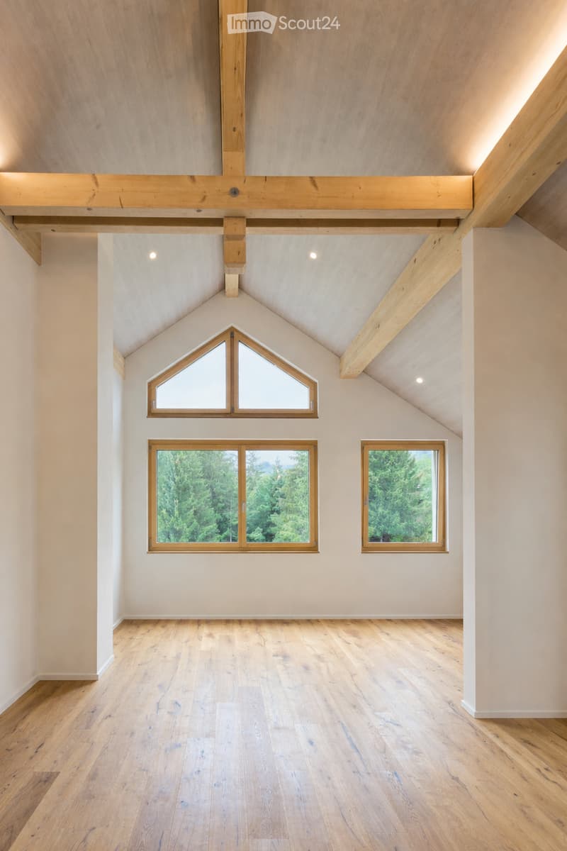 An empty attic space with exposed wooden beams, wooden flooring, and large windows offering a view of greenery outside.