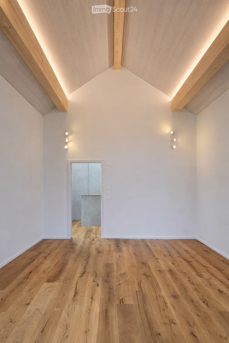 Empty room with light wooden flooring, white walls, and recessed lighting in the ceiling. A doorway leads to another room with a visible railing.