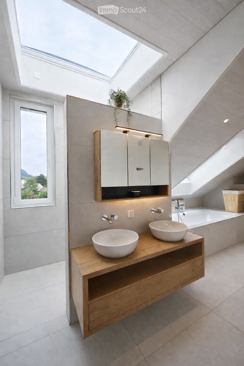Modern bathroom with two sinks on a wooden vanity, a large mirror with built-in cabinets, a bathtub, and a skylight window providing natural light.