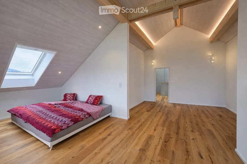 Modern bedroom with wooden flooring, slanted wooden ceiling, skylight window, and a neatly made bed with pillows and a colorful blanket.