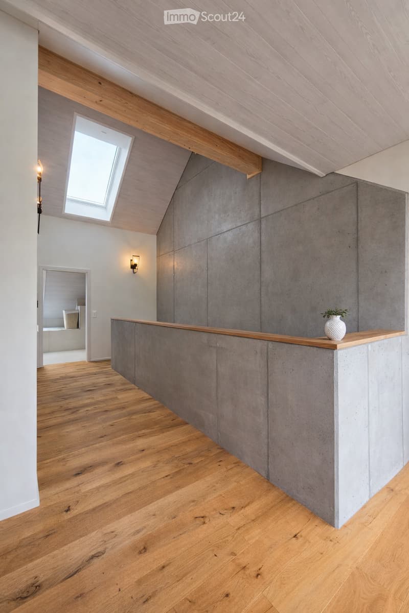 Modern, minimalist room with wooden flooring, exposed wooden beams, a concrete counter, and a small vase on the counter. A skylight provides natural light, and there are wall-mounted lamps for additional illumination.