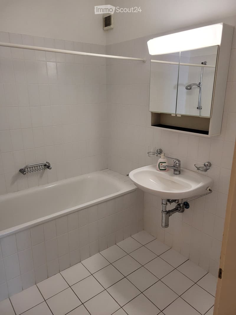 Bathroom with white tiled walls and floor, featuring a bathtub, a sink with a faucet, a mirror cabinet, and a soap dispenser.