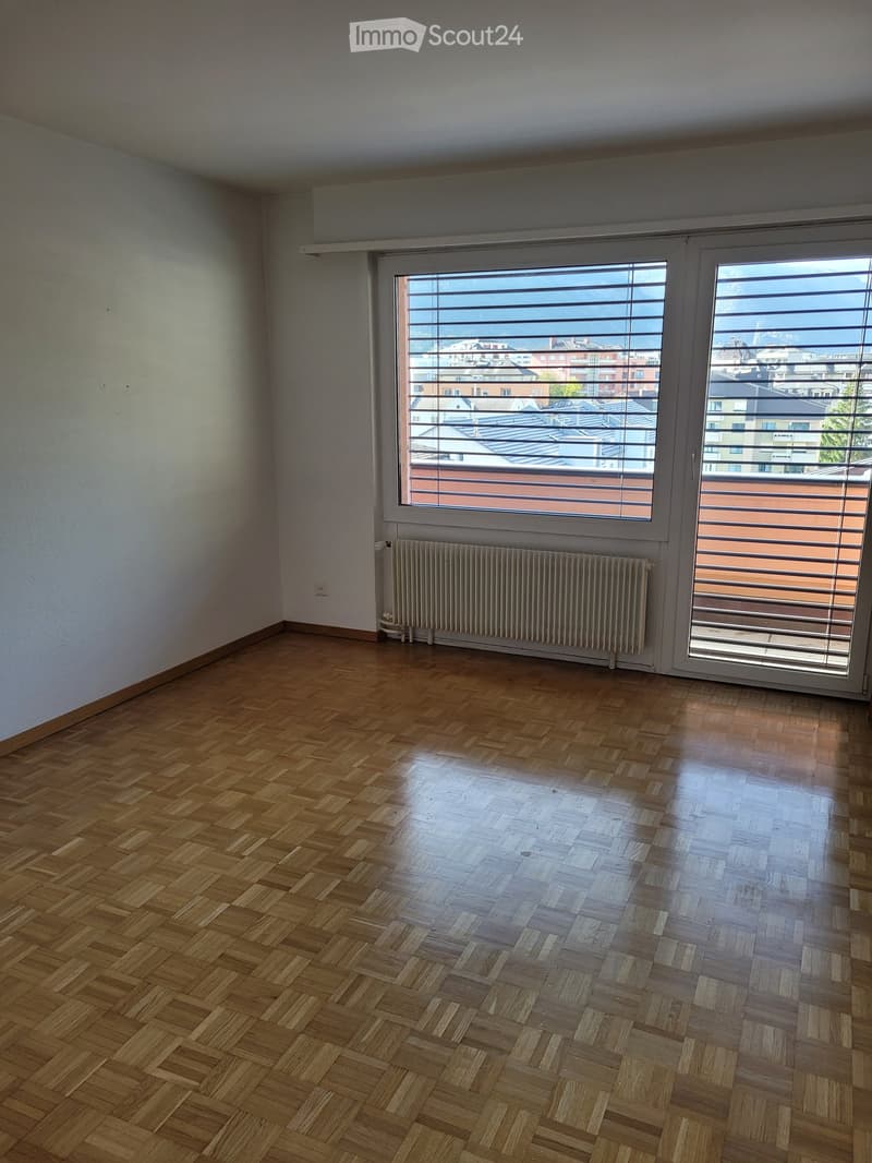 Empty room with wooden herringbone flooring, white walls, large window with blinds, radiator, and sliding glass door leading to a balcony.