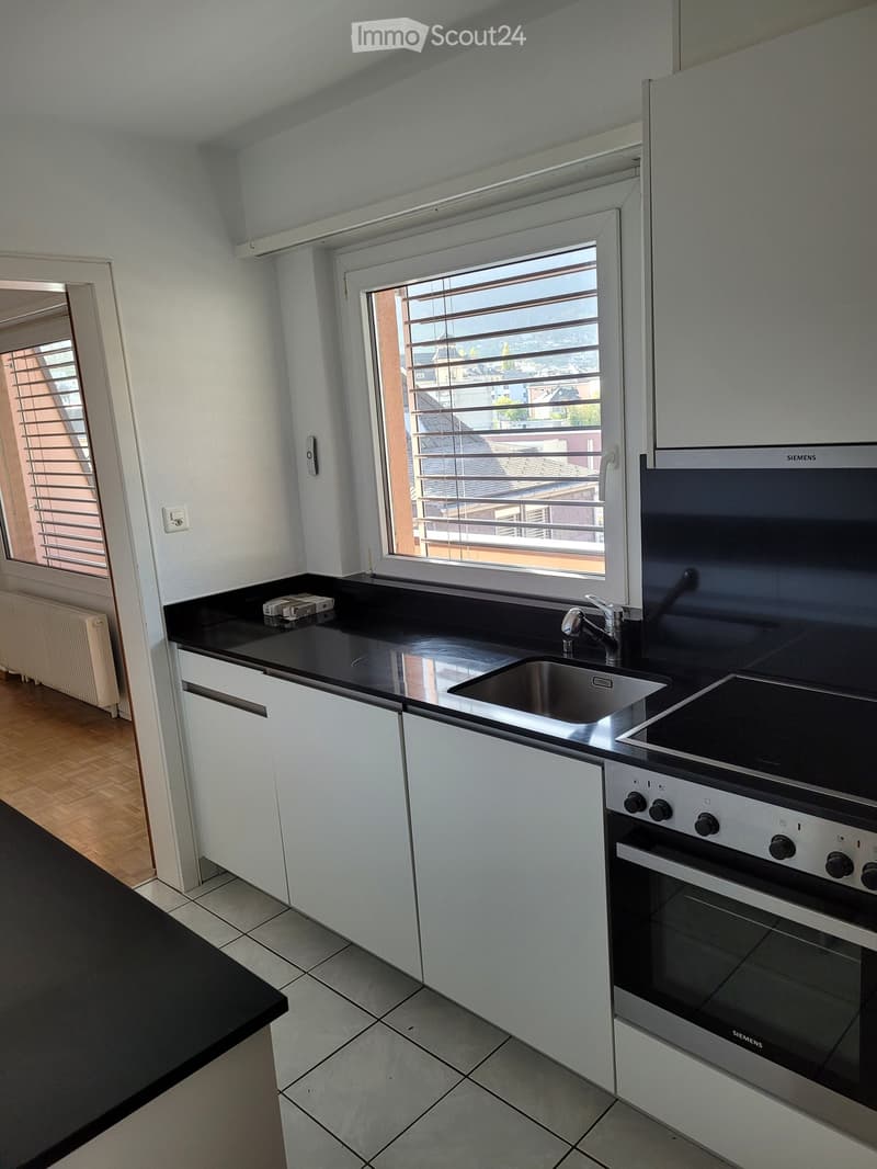 Modern kitchen with white cabinets, black countertop, stainless steel sink, and Siemens oven. Features large windows with blinds and a view of the city.