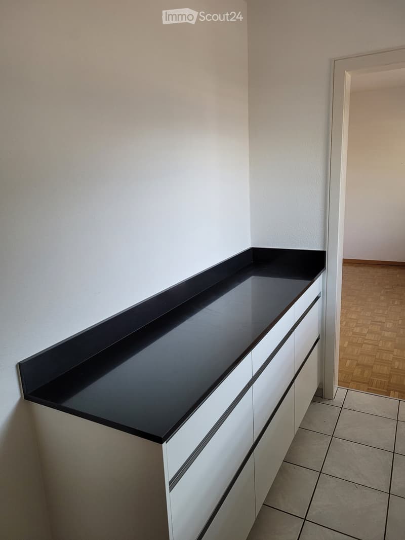 Empty room with white walls, tiled floor, and a black countertop with white cabinets attached to the wall. Wooden flooring is visible in the adjacent area.