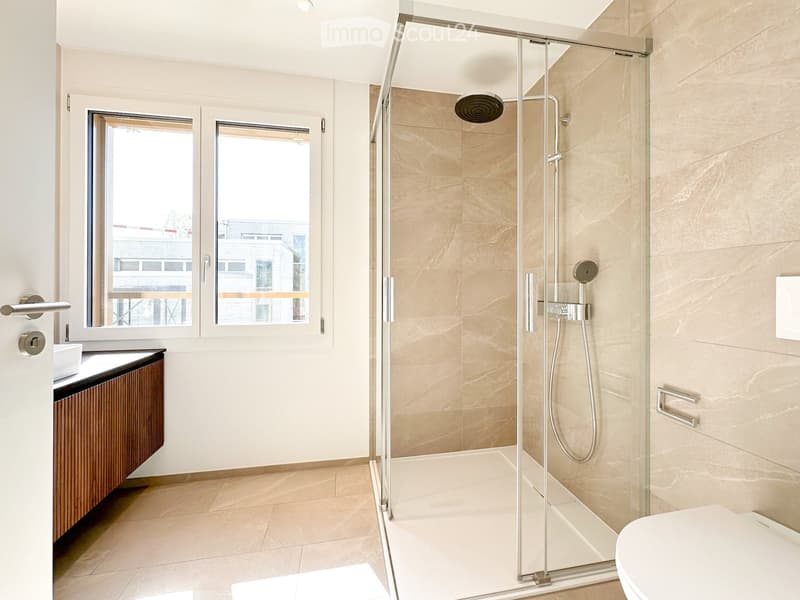 Modern bathroom with a glass-enclosed shower, a toilet, a sink with a wooden cabinet, and a large window offering a view of the city.