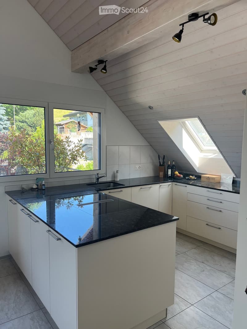 Modern kitchen with white cabinets, black countertops, and a large window. Features include a sink, faucet, wine bottles, fruit basket, and cutting board on the counter.