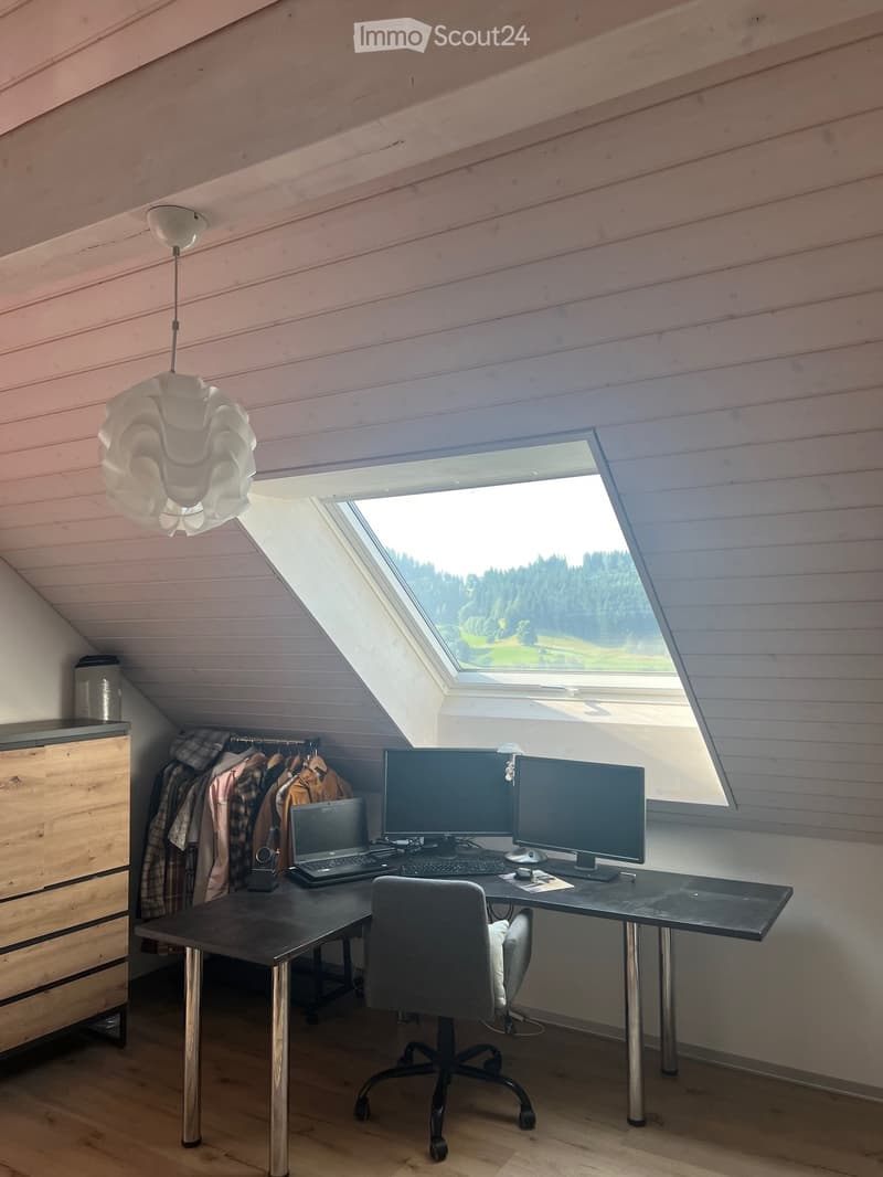 Bright attic workspace with slanted wooden ceilings, large skylight window offering a scenic view, modern desk setup with dual monitors, laptop, keyboard, mouse, ergonomic chair, and a stylish hanging lamp.