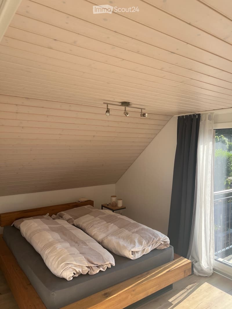 Cozy bedroom with a wooden bed, nightstand, and large window with curtains. The room features a slanted wooden ceiling and warm lighting.