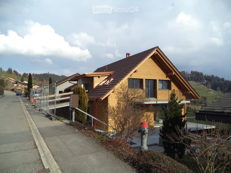 Two-story house with a wooden facade, dark brown roof, and large windows. The house features a balcony and is situated in a rural area with greenery and hills in the background.