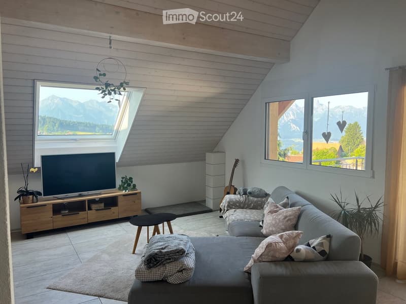 Modern living room with large windows offering a scenic mountain view, featuring a grey sectional sofa, coffee table, TV, guitar, and decorative elements.