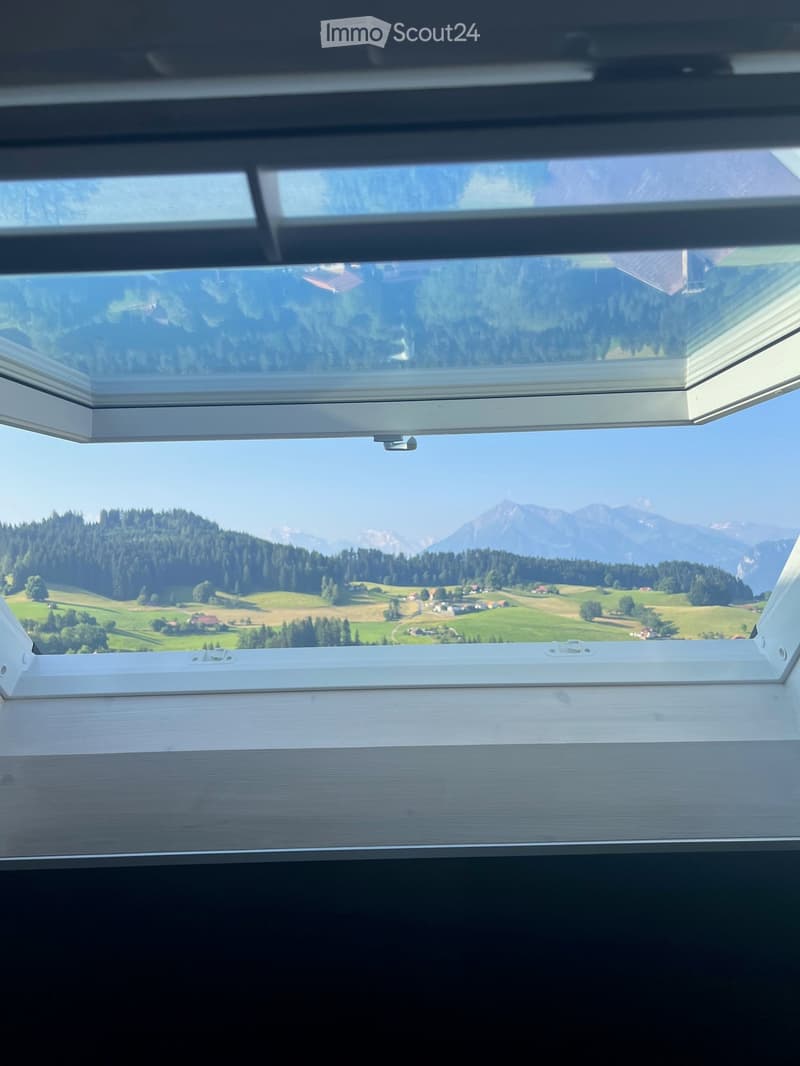 A panoramic view from a balcony or terrace showcasing a lush green valley, scattered houses, and distant mountains under a clear blue sky.