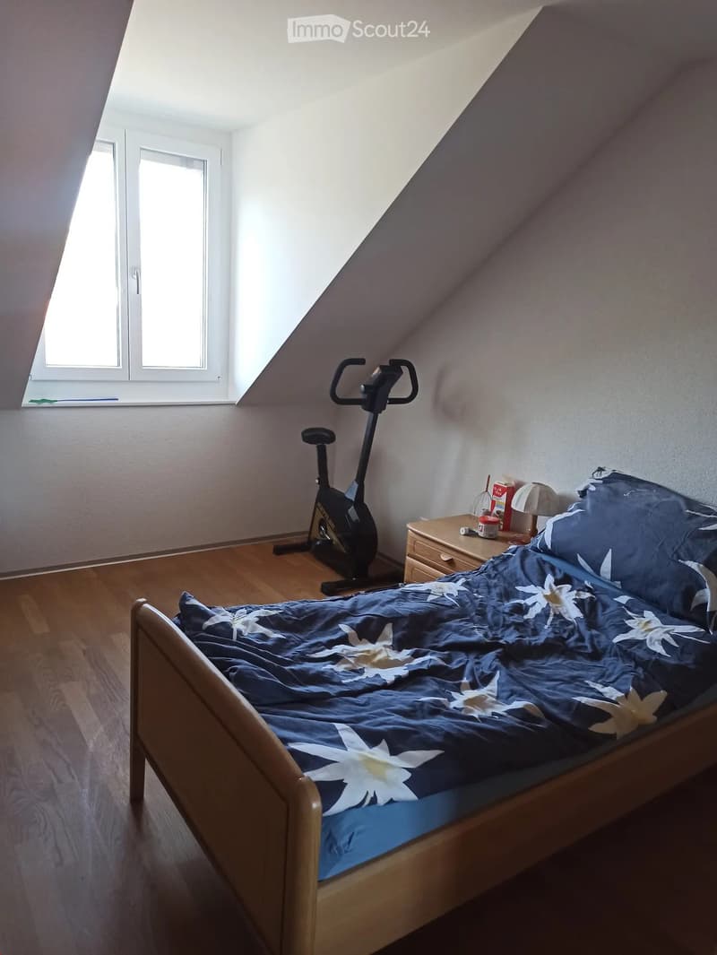 Cozy attic bedroom with wooden floor, slanted ceiling, large skylight, bed with blue floral duvet, nightstand with lamp, and exercise bike in corner.