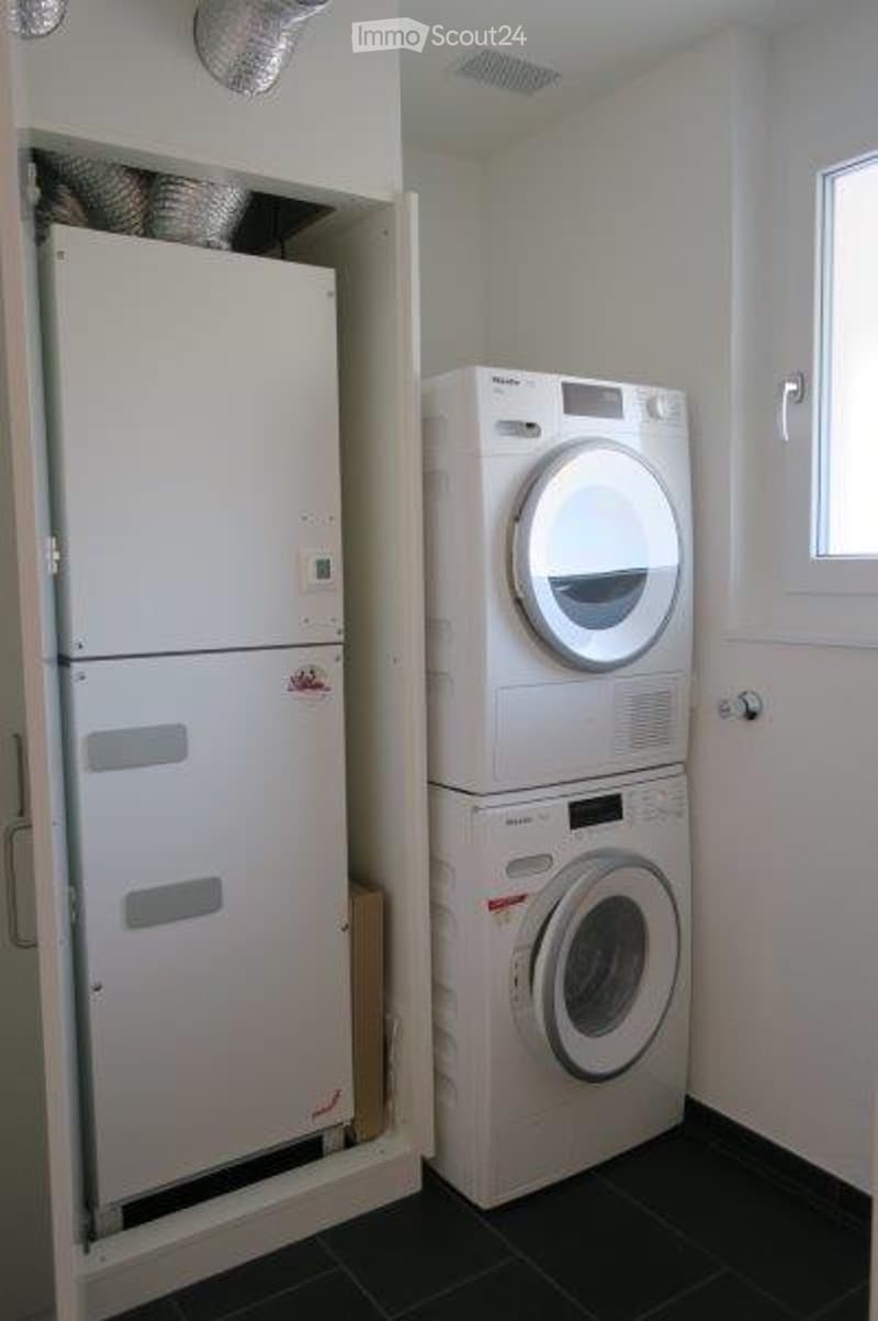 Compact bathroom with a stacked washer and dryer unit and a small refrigerator, white walls, dark tiled floor.