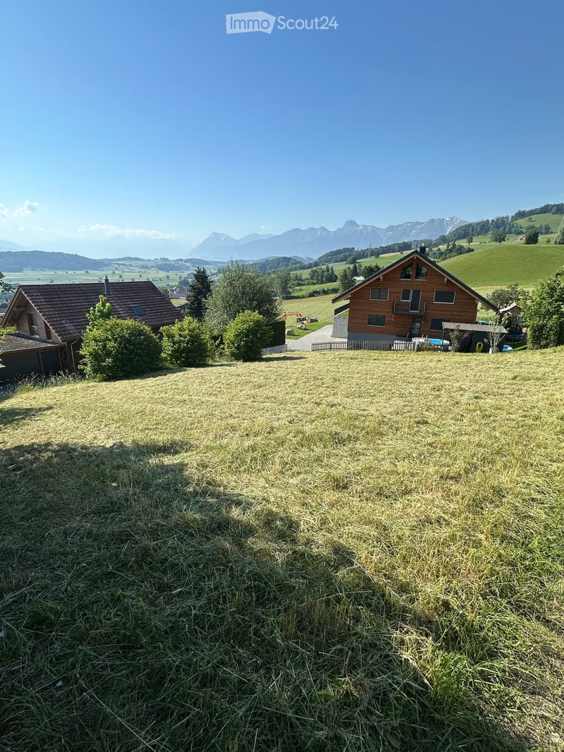 Two-story wooden house with a red-tiled roof, surrounded by green fields and mountains in the background.