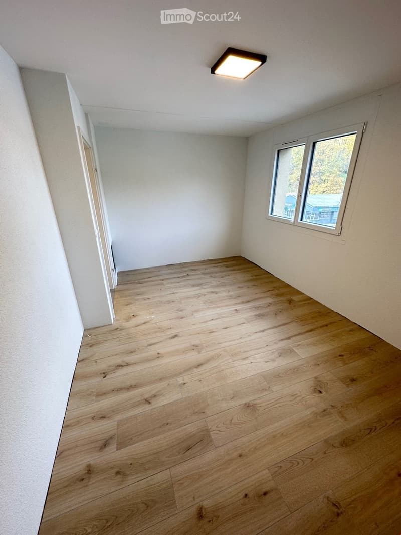 Empty room with light wooden flooring, white walls, and a single window letting in natural light. The room has a square layout with a door visible on one side.