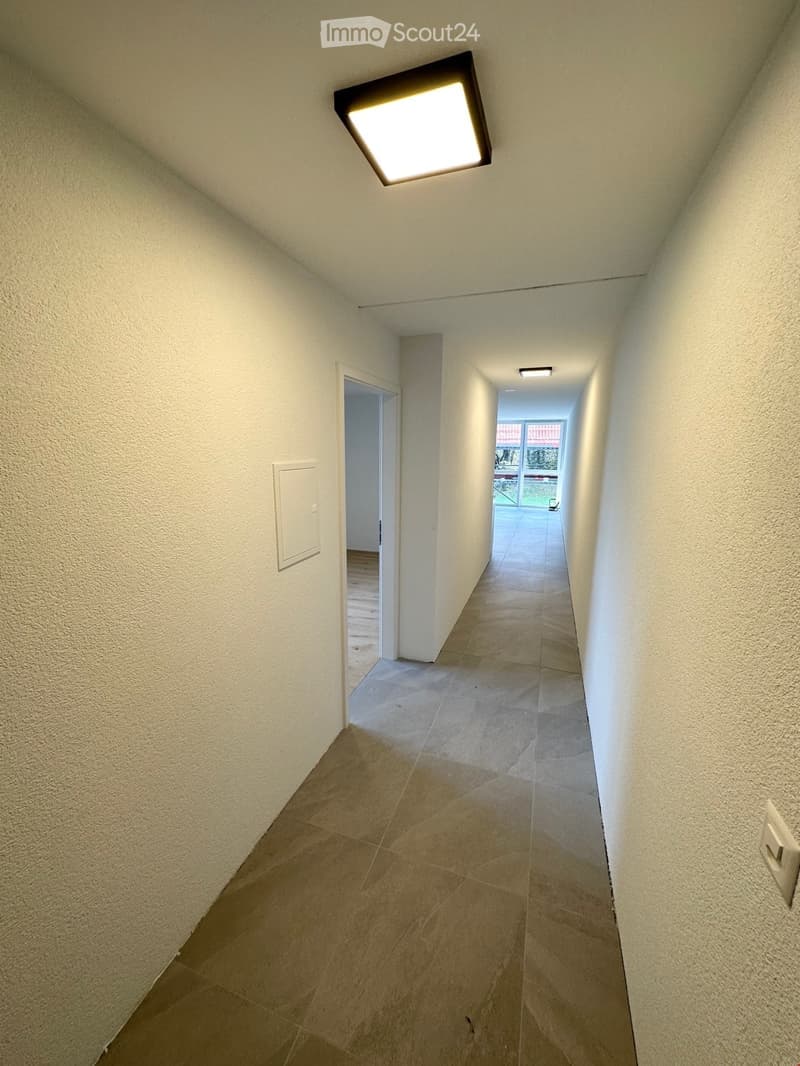 Empty hallway with beige tiled flooring, white walls, and recessed lighting. A door leads to another room at the end.