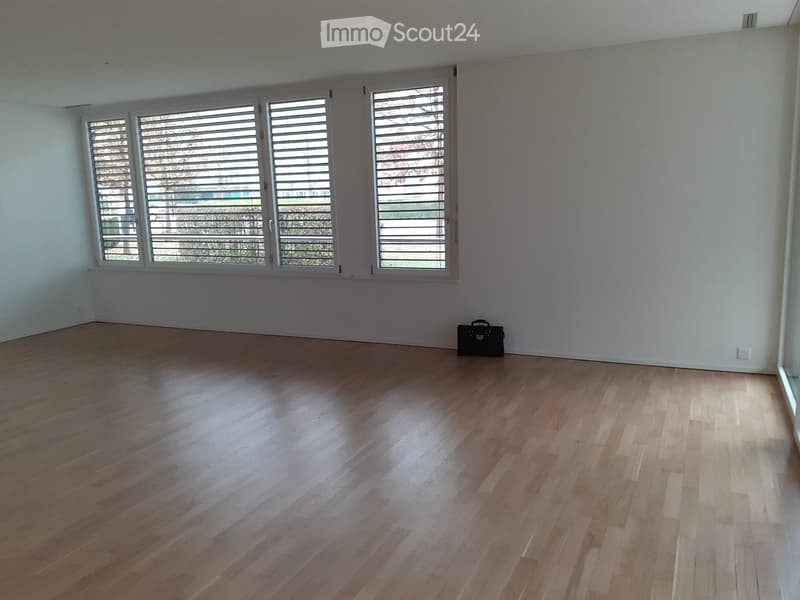 Empty room with light wooden flooring and large windows with blinds. A small black bag is placed against the wall.