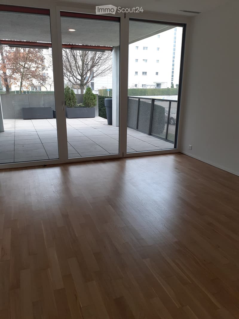 Empty room with wooden flooring and large glass sliding doors leading to a balcony area with planters and a view of a building.