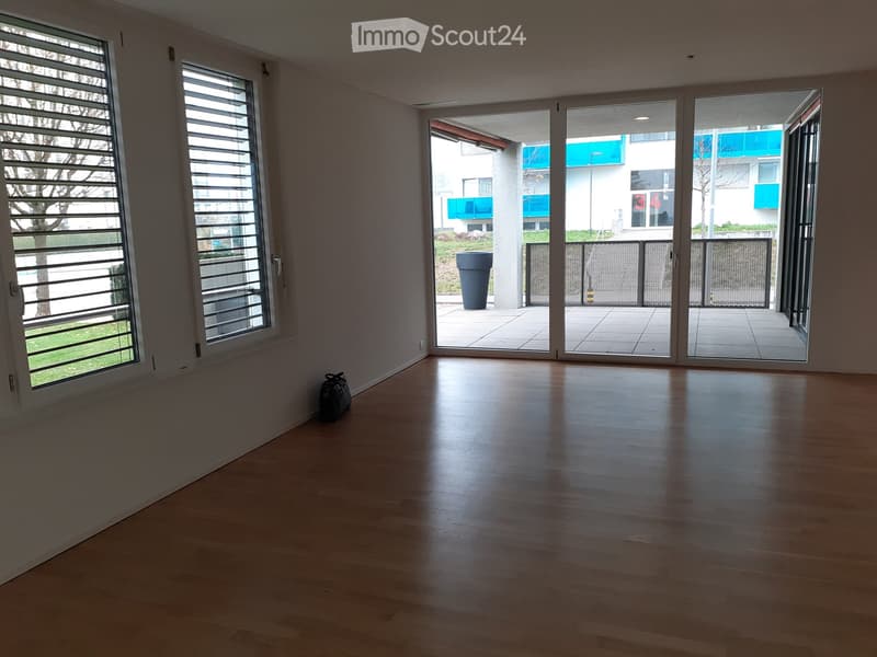 Empty room with wooden flooring, large windows with blinds, and sliding glass doors leading to a balcony area.